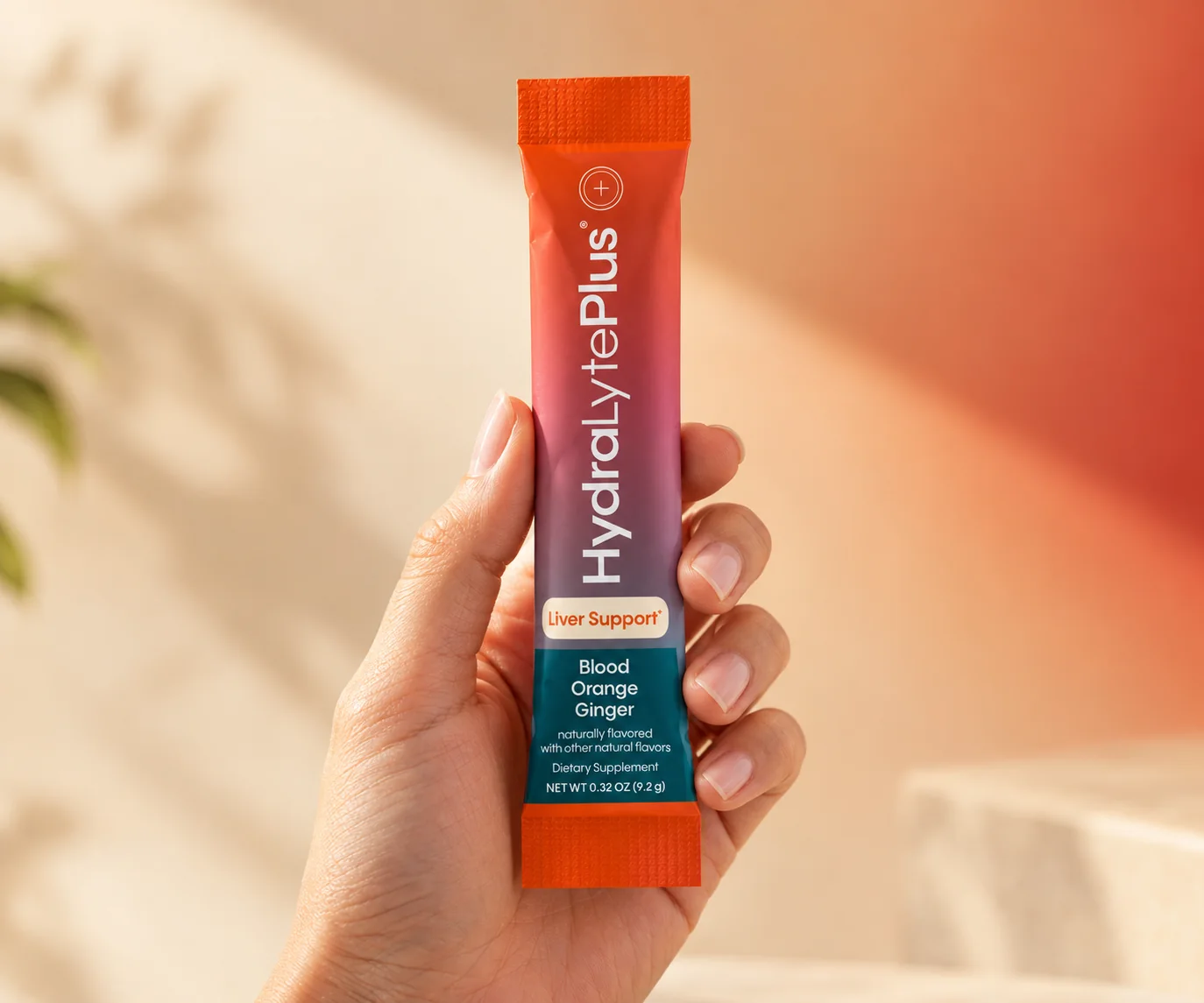 A hand holding a HydraLytePlus stick pack against a warm wall