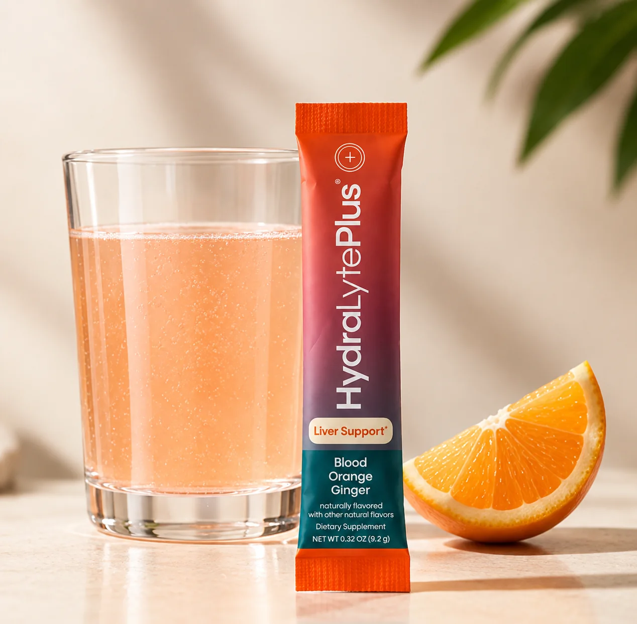 A HydraLytePlus stick pack beside a glass of blood orange drink and an orange slice